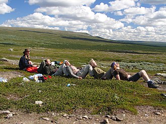 6 Personen sitzen hintereinander auf einer weitläufigen Tundra (jeweils an die aufgestellten Schienbeine der hinteren Person gelehnt) und genießen die Sonne
