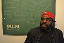 Hanif Willis-Abdurraqib pictured in front of a map of Ohio. He wears a black shirt and jacket and a red baseball cap, turned backwards, and looks off-camera.