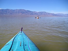 Un uomo che fa kayak in un lago marrone, montagne sullo sfondo, un kayak in primo piano