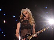 Picture of a middle-aged blond woman uptill the waist, singing in front of a microphone. Her hair is in waves and falls on her shoulders. She wears a black bra covered with net and wears a white hat on her head. She plays an electric guitar. Behind her, to her left, a flood light is visible.