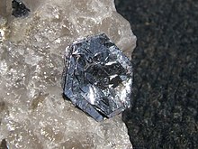 Glänzende, silbrige, flache, sechseckige Kristalle in etwa parallelen Schichten sitzen blütenartig auf einem rauen, durchscheinenden kristallinen Quarzstück.