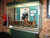 A glass display case containing a large brass model of a steam locomotive. At the back of the case is a black and white photograph of a man.