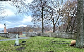 Links im Bild ist der See zu sehen, ebenfalls die auf einem sehr hohen Podest stehende Skulptur. Rechts im Bild befinden sich Bäume, an deren Fuß die Gedenkkugel liegt.