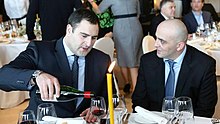 Man pouring wine at a candlelit table with a fellow diner. The ambience of the room can be described as warm with people mingling in the background.