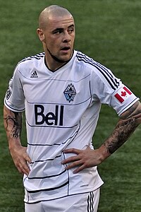 A man wearing a white T-shirt and white shorts, standing on a soccer field with his hands at on his hips, looking to his left.