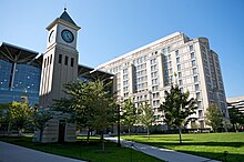 Zwei moderne Glas- und Betongebäude nebeneinander vor einer offenen Rasenfläche mit einem kurzen Uhrenturm auf der linken Seite.