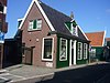 Grotendeels houten huis met zadeldak parallel aan straat. Ingang met bovenlicht tussen pilasters. Het gesneden fries tussen de pilasterkapitelen met emblemen van de koopvaardij