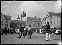 Young girls shooting a ball into a Wicker basketball. Image is in black and white. Girls are wearing skirt. A male, pant wearing umpire stands on the court.