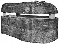 Black and white image of a dark stone coffin viewed laterally, the coffin lies on the ground, the trough and lid are separated with wedges.