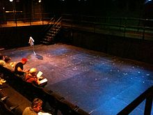 A sunken black box theater space with several audiencemembers in the first two rows of chairs facing the stage