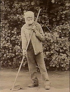 old bearded mat wearing trousers and a coat, leaning on a pole