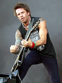 Rob Holliday wearing dark pants and a black sleeveless print shirt, playing electric guitar onstage