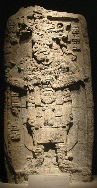 A relief sculpture showing a richly dressed human figure facing to the left with legs slightly spread. The arms are bent at the elbow with hands raised to chest height. Short vertical columns of hieroglyphs are positioned either side of the head, with another column at bottom left.
