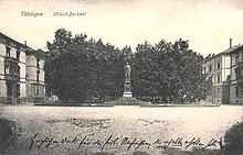 Photo en noire et blanc d'une place. Au fond de celle-ci se trouve une statue d'homme et des arbres.