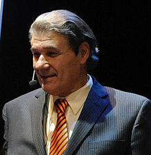 Photographie en couleur. Morales est représenté en buste avec une lumière venant de sa droite. Il porte un costume et une cravate rayés et porte un micro et une oreillette.