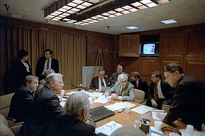 Des hommes en costume assis autour d'une table ovale discutent. Des papiers sont sur la table. Au fond, une télévision des années 1980 est allumée.