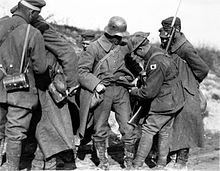 Black and white photo of a group of seven men standing while wearing military uniforms. One of the men, armed with a rifle, is going through the pockets of another man