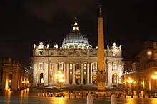 Behind a large monolithic obelisk, the facade of St. Peter's Basilica, lit by floodlights, rising majestically against the night sky