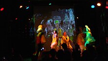 Enthusiastic Indian dancers wear brightly colored clothing under colorful lights on a dark stage, in front of artistic video projected onto a screen. The photo is noisy, with lots of motion blur due to a longer exposure in the dark club, but you can make out a couple faces.