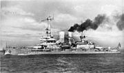 A large warship, thick black smoke pouring out of its rear funnel, steams through the calm sea