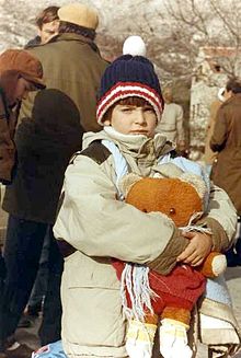 Một đứa trẻ mặc quần áo mùa đông và ôm một con gấu bông lớn nhìn vào máy ảnh, với những người lớn ăn mặc tương tự khác đang bận rộn ở phía sau