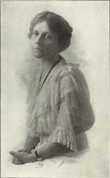  A woman seated with cropped or pulled back hair, wearing a white lacy dress, dark banded watch and dark ribbon or necklace. She faces the viewer.