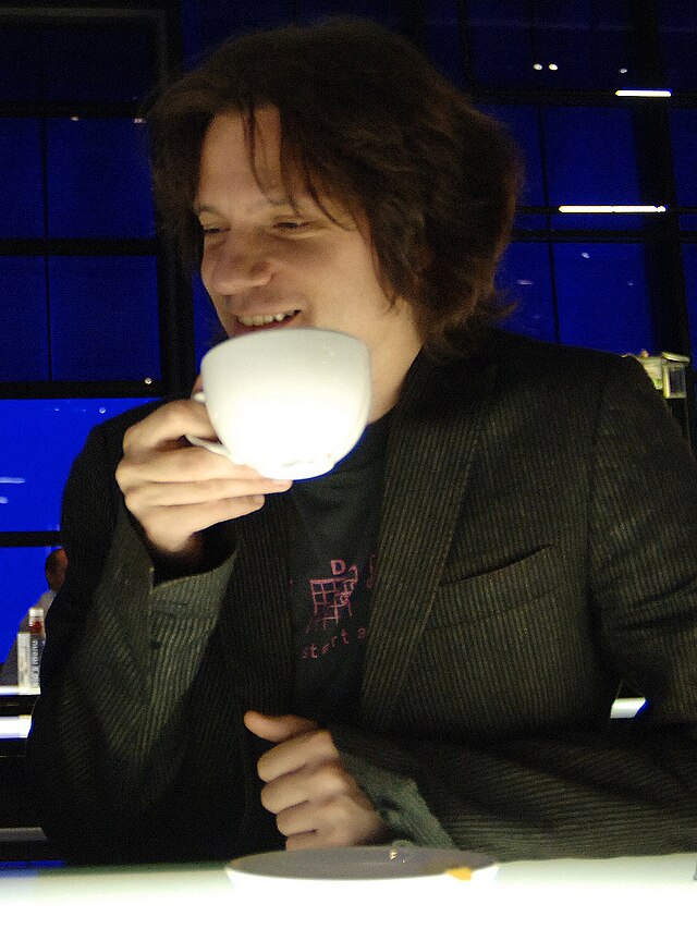 Guy Sigsworth looking towards his right and smiling, while drinking from a cup in his right hand.