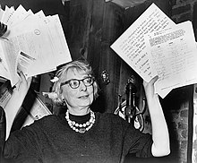 Woman in glasses waving documents in air.