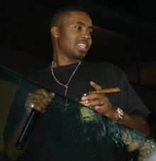 Nas holding a cigar, a glass, and a microphone. He is looking away from the camera.