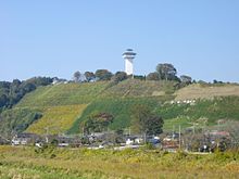 Kleurenfoto van een heuvel bedekt met groen, met een dorp aan de voet en een witte toren bovenaan, tegen een blauwe lucht.