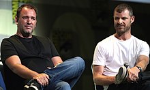 Two adult males sitting in chairs with their left legs crossed.