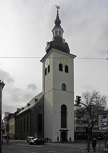 Weiß verputzter Kirchturm mit Rundbogenfenstern und einer schiefergedeckten Zwiebelhaube, dahinter anschließend ein einfaches Kirchenschiff aus Bruchstein mit hohen Rundbogenfenstern.