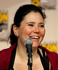 A caucasian woman with black hair tied back, smiling into a microphone, with a vague symbol behind her.