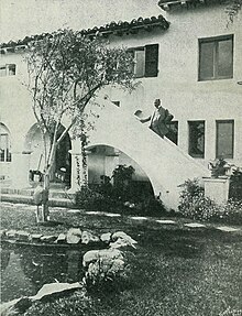 Photo en noir et blanc d'un homme debout sur les escaliers extérieurs d'une maison de type villa espagnole.