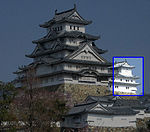 Une petite tour de château à trois étages à côté d'une grande tour à cinq étages.  Les deux ont des murs blancs, des toits sombres et sont construits sur une plate-forme de pierres brutes.