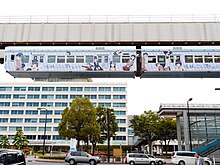 Foto colorida de um monotrilho suspenso passando sobre carros, em uma cidade, sob um céu leitoso.