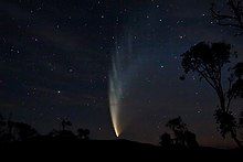 Num céu escuro, com alguma estrelas, o núcleo brilhante do cometa está próximo ao horizonte e sua calda, incialmente amarelada, se estende para o alto e para a direita mudando para a coloração azul e ficando cada vez mais extensa.