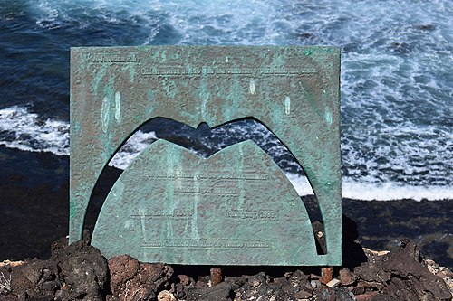 Uma placa de metal com aspecto esverdeado e corroído sob pedras de uma linha costeira e frente ao oceano. Tem a forma de uma gaivota vasada e leva dizereres em espanhol, descritos na leganda.