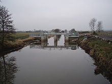 Gerader Wasserlauf, umgeben von Feldern und durchzogen von Metall- und Betonkonstruktionen.