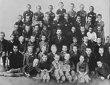 Foto di classe in bianco e nero di cinquanta ragazzi riuniti attorno al loro maestro di scuola;  il giovane Hitler è in piedi al centro della prima fila.