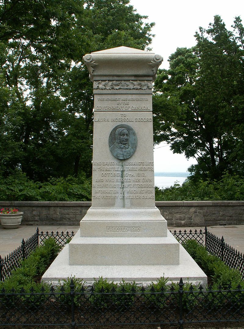 An upright stone monument surrounded by a low hedge. In the middle is an upright oval of an elderly woman's face in relief.