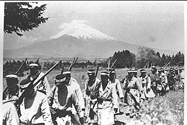 Groupe d'hommes en uniforme militaire et fusil à l'épaule marchant le long d'un chemin. Le mont Fuji est visible au loin.