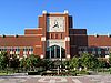 The front of an athletic facility with dominating red brick and an analog clock that extends high in the middle with trees and a long narrow pool in the foreground.