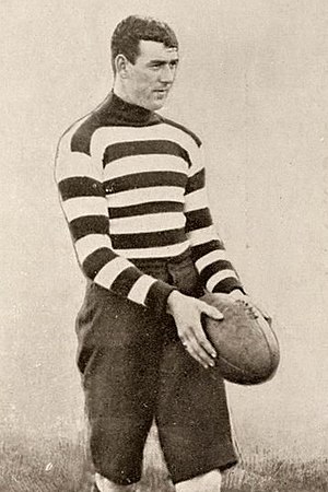 A dark-haired footballer in a long-sleeve horizontally-striped guernsey holding a football
