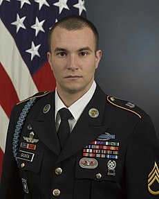 Head and torso portrait of a young white man in a formal military uniform with a U.S. flag in the background