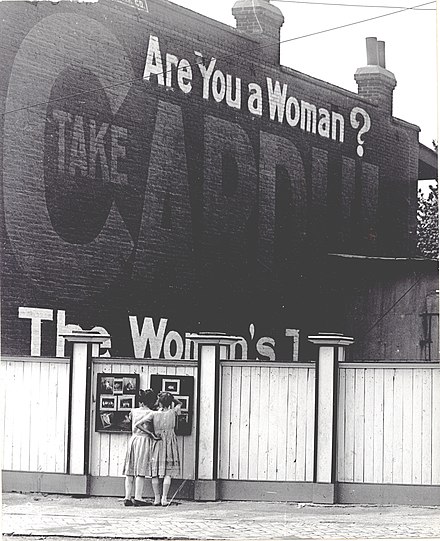 Two girls examining a bulletin board posted on a fence. An advertisement painted above them asks "Are You a Woman?".