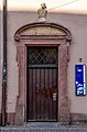 An old wooden door surrounded by a sandstone wall. A blue university of freiburg plaque is on the wall next to it, indicating it is a university building. The door appears accessible, though heavy to open.