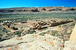 Large circular depression outlined by a stone wall. The bottom is flat and grassy, and has a collection of rectangular stone foundations and smaller circles of stone. A great sandstone cliff towers in the background, and beneath the cliff are other stone foundations that are larger and higher.