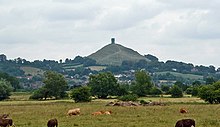 A mound surmounted by a tower in the distance. In the foreground are fields with cows and small trees and bushes.