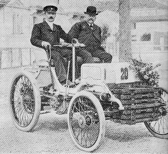 Photo de deux hommes assis dans une voiture d'apparence hippomobile avec un imposant radiateur à l'avant.